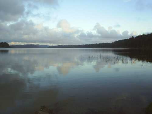 A calm lake reflecting clouds and trees, surrounded by a serene landscape under a cloudy sky.