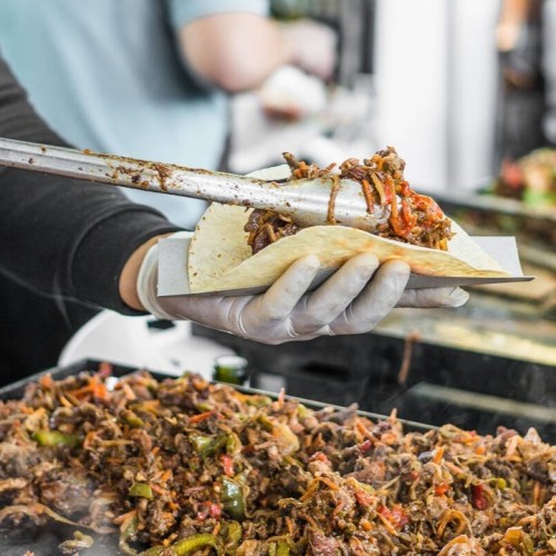 A hand holding a taco filled with seasoned meat and vegetables, with a grill of sizzling meat in the background.