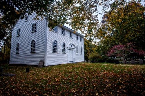 A white historic building surrounded by autumn leaves and trees, with arched windows and a grassy area.