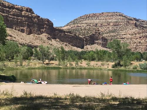 A serene landscape featuring a calm pond surrounded by trees and rocky hills, with people enjoying the water.