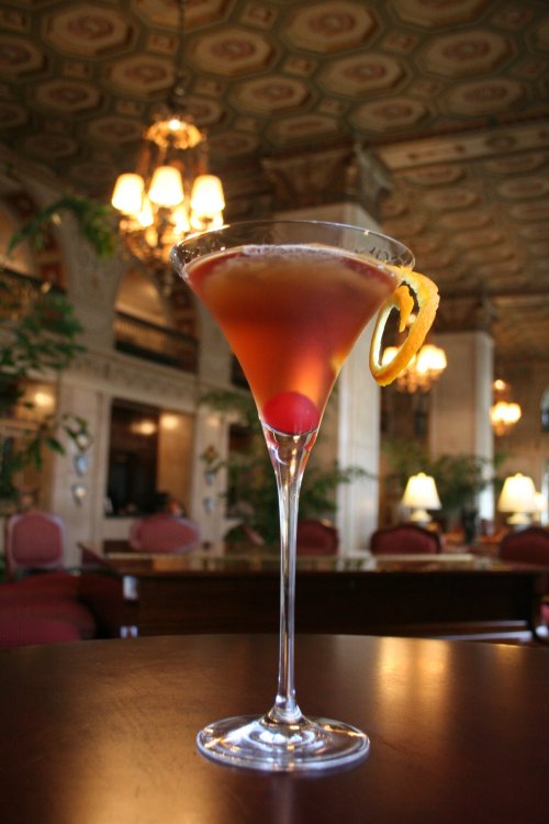 A cocktail in a tall glass garnished with a cherry and orange slice, set against an elegant, vintage interior.