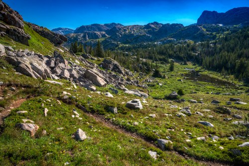 A scenic mountain landscape featuring rocky terrain, lush greenery, and distant peaks under a clear blue sky.