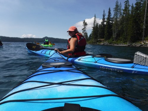 A person in a kayak on a calm lake, surrounded by trees and another kayak in the background under a clear sky.