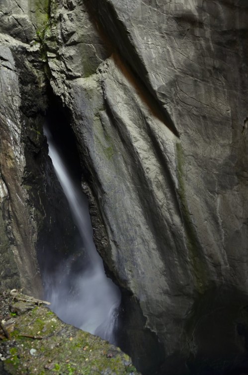 A narrow canyon with a waterfall cascading down rocky walls, surrounded by lush greenery.