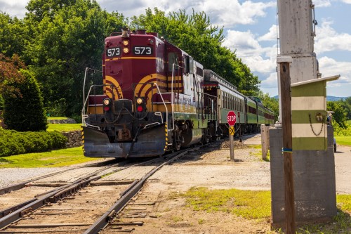 A vintage train with a red and yellow locomotive and green passenger cars approaches a stop sign on a sunny day.