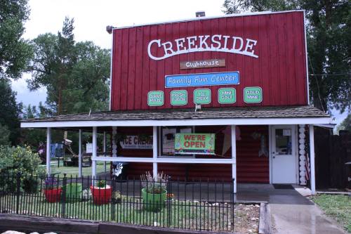 Red wooden building with "Creekside Clubhouse" sign, open for business, surrounded by greenery and colorful planters.