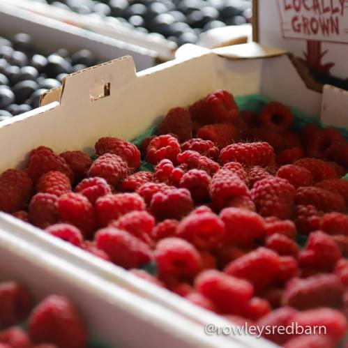 A close-up of fresh raspberries in a market display, with blueberries visible in the background.