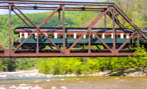 A vintage train crosses a metal bridge over a river, surrounded by lush green trees and hills.