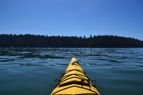 A yellow kayak on calm water, with a forested shoreline in the distance under a clear blue sky.