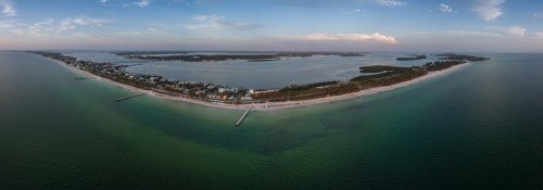 Aerial view of a coastal area with sandy beaches, water, and a small town along the shoreline under a blue sky.