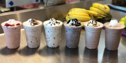 Six milkshakes in cups, topped with whipped cream and various toppings, lined up on a counter with bananas in the background.