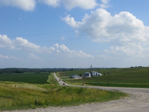 A rural landscape with a winding road, green fields, and a farm with silos under a blue sky with fluffy clouds.