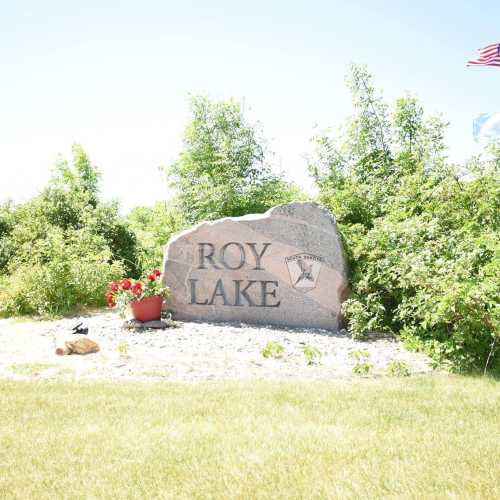 A stone sign reading "Roy Lake" surrounded by greenery and grass under a clear blue sky.