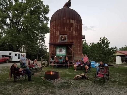A group of people gathered around a fire pit near a rustic silo, enjoying an outdoor gathering at dusk.