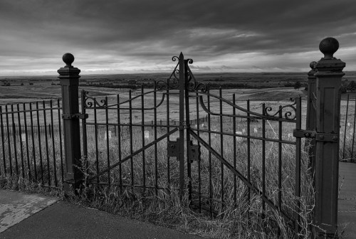 A wrought iron gate stands open, revealing a vast landscape under a dramatic, cloudy sky.