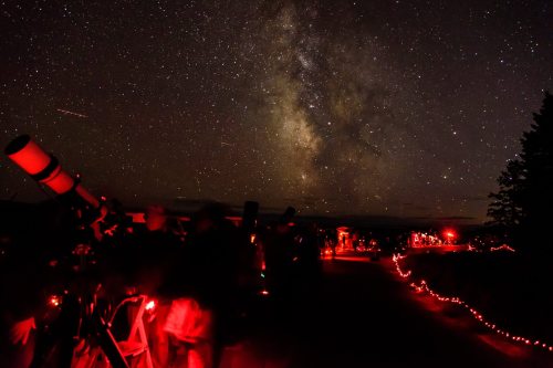 A stargazing event with telescopes set up under a starry sky, featuring the Milky Way and red lights illuminating the scene.