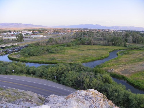 Aerial view of a winding river surrounded by greenery, with a town and mountains in the background at dusk.