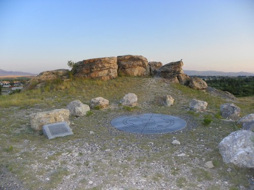 A rocky hilltop with a circular metal plaque and informational sign, surrounded by grass and distant mountains.