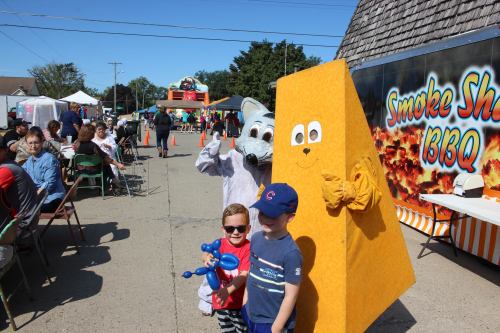 Two children pose with a mascot in a bright yellow costume at a festive outdoor event, with food stalls in the background.