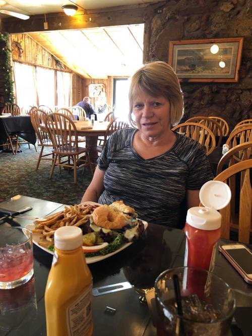 A woman sits at a table in a restaurant, smiling with a plate of food, including a burger and fries, in front of her.