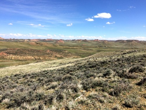 A vast, open landscape with rolling hills, sparse vegetation, and a clear blue sky dotted with clouds.