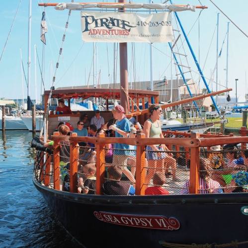 A pirate-themed boat with passengers enjoying a sunny day at the harbor, surrounded by sailboats and clear blue skies.