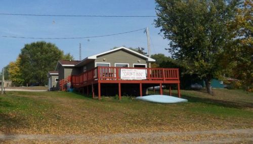 A small house with a red deck and sign, surrounded by trees and autumn leaves, on a sunny day.