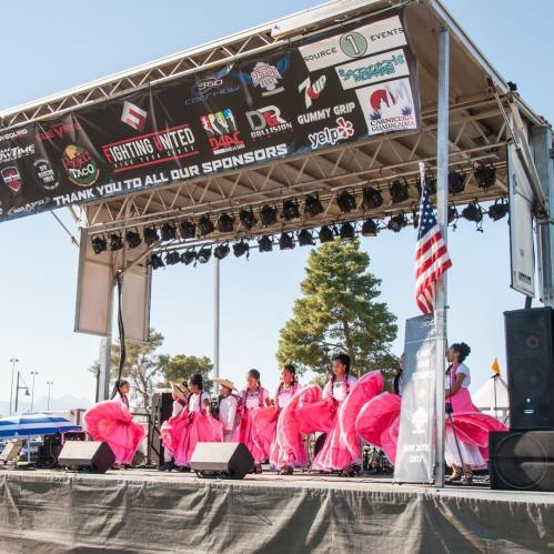 A group of dancers in pink dresses perform on stage under a banner thanking sponsors at an outdoor event.