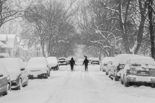 Two people walk down a snowy street lined with parked cars, surrounded by trees and heavy snowfall.