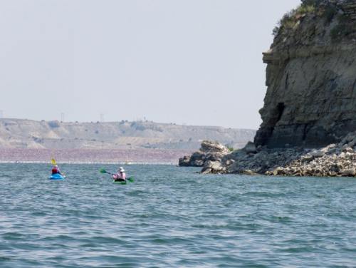 Two kayakers paddle on a calm lake near a rocky cliff, with distant hills visible in the background.