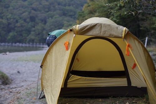 A yellow camping tent set up by a lakeside, with a person adjusting it in the background among trees.