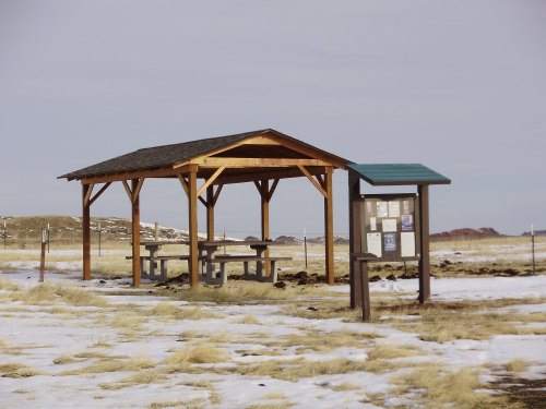 A wooden picnic shelter with tables, surrounded by snow and grass, near a bulletin board in a rural area.