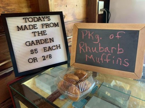 A sign displays "Today's made from the garden $5 each or 2 for $8" next to a chalkboard listing "Pkg. of Rhubarb Muffins."