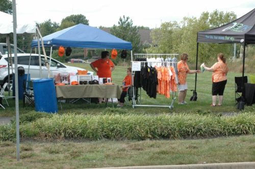 A vendor booth with orange shirts and hats, featuring people interacting under tents in a grassy area.