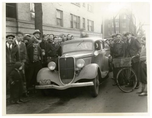 A crowd of people gathers around a vintage car on a city street, with a bicycle parked nearby.