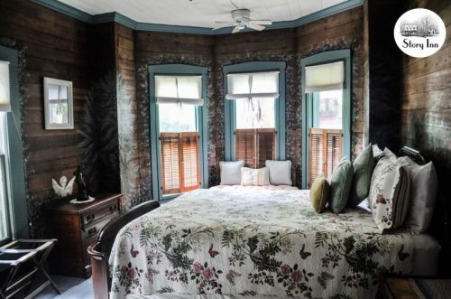 Cozy bedroom with a floral quilt, wooden walls, and large windows, featuring a vintage dresser and soft lighting.