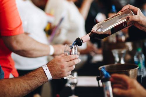 A hand pours red wine into a glass at a tasting event, with people mingling in the background.
