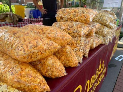 Bags of colorful popcorn stacked on a table at a market, with a checkered tablecloth in the background.