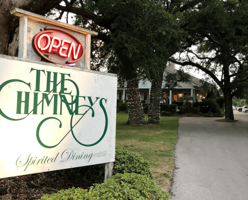 A sign for "The Chimneys" restaurant with an "OPEN" neon light, surrounded by trees and a well-lit pathway.