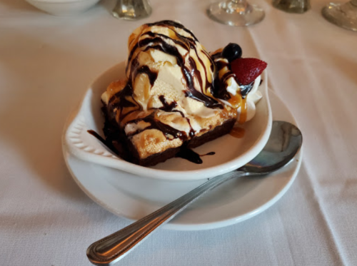 A dessert dish featuring a brownie topped with vanilla ice cream, chocolate sauce, and a cherry, served on a white plate.