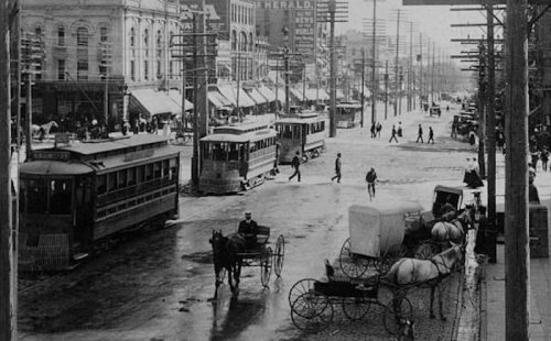 Historic street scene with trams, horse-drawn carriages, and pedestrians on a snowy day. Black and white photo.