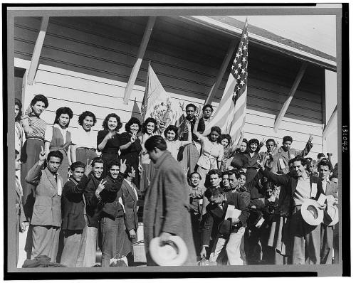 A large group of young people, some holding flags, poses together on a building's steps, celebrating with smiles and gestures.