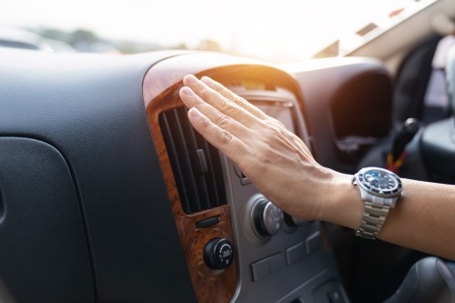 A hand reaching towards a car's dashboard, adjusting the air vent with sunlight in the background.