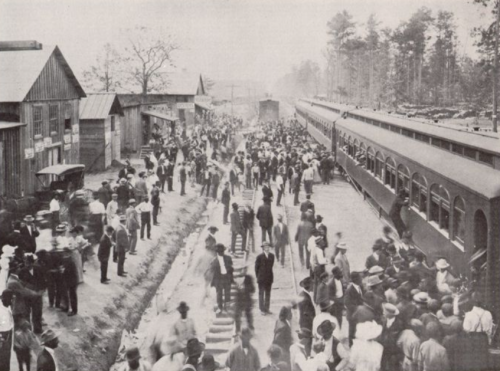 A crowded train station with people gathered around, vintage buildings in the background, and trains on the tracks.