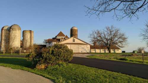 A scenic farm with silos, a large house, and a tree, set against a clear blue sky and green grass.