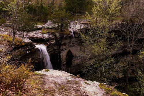 A serene waterfall cascades over rocky cliffs, surrounded by lush greenery and trees in a tranquil natural setting.
