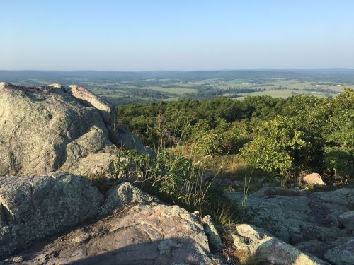 A scenic view from a rocky outcrop, overlooking lush green valleys and distant hills under a clear blue sky.