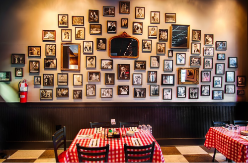 A restaurant interior featuring a wall adorned with numerous black-and-white framed photos and checkered tablecloths.