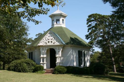 A charming octagonal building with a green roof, decorative trim, and surrounded by lush greenery and trees.