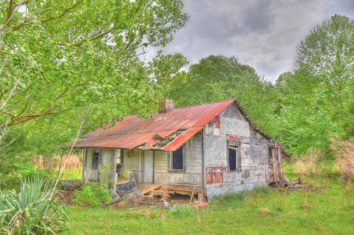 Abandoned house with a rusty roof, surrounded by overgrown greenery and trees under a cloudy sky.